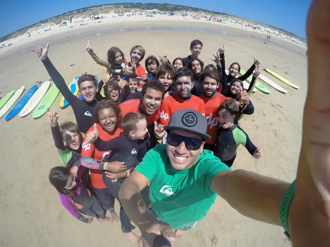 Grande grupo de crianças e treinadores a sorrir e a fazer o gesto do shaka na praia, junto às pranchas
